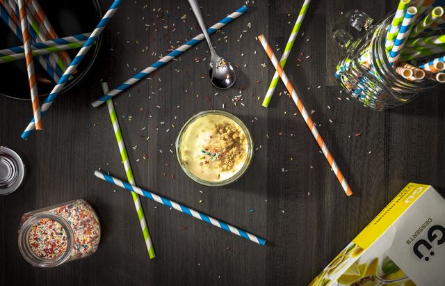 Colorful sprinkles scattered on dark wooden table, with a glass of dessert and striped paper straws positioned randomly. Spoon and jar with additional sprinkles visible. Great for illustrating a festive or party theme, beverage or dessert promotions, and DIY craft blogs.