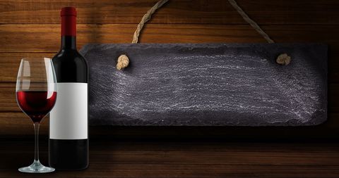 Sophisticated scene features wine bottle and glass on a wooden surface with a blank slate sign. Perfect for use in promotional materials for wine tastings, business celebrations, or events catering to viniculture enthusiasts. Effective in blogs about wine pairings or lifestyle articles.