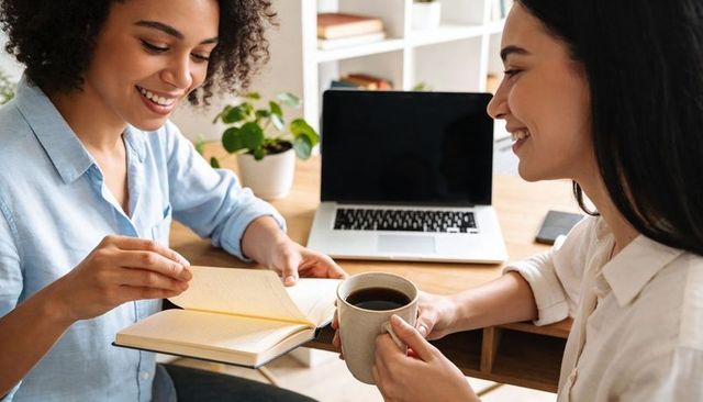 Smiling colleagues flipping notebook pages while sharing coffee at sunlit home workspace