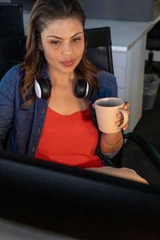 Focused woman working at desk sipping coffee with headphones and computer monitor