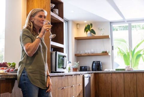 Mature woman sipping wine while leaning on wooden countertop in bright modern kitchen
