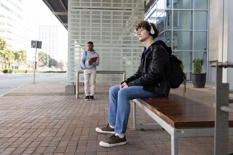 Diverse teen students waiting at urban shelter listening to music with backpacks