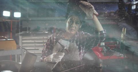 Female welder fabricating metal at workbench wearing plaid shirt, apron and gloves