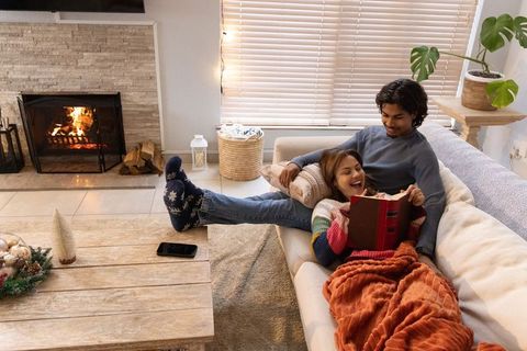 Multicultural couple relaxing on sofa reading by fireplace with cozy blanket and decor