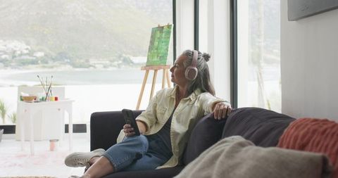 Middle-aged woman relaxing with headphones and smartphone on sofa beside easel with waterfront view