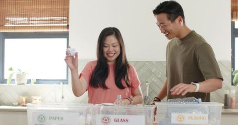Asian couple sorting recyclables in bright kitchen with labeled paper glass plastic bins