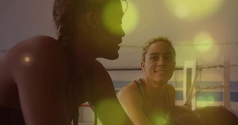 Two women resting and smiling after boxing training inside gym with warm lens flare