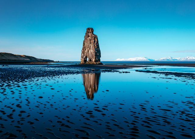 Majestic solitary rock formation stands tall amid calm waters during low tide, reflecting clearly against the serene blue sky. The scene captures the rugged beauty of a coastal landscape. Perfect for nature magazines, travel brochures, websites, and educational materials focusing on geology and coastal environments.