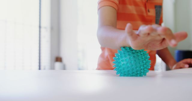 This image features a young child wearing an orange shirt playing with a spiky sensory ball on a white table in a bright room. Perfect for educational content, early childhood development materials, sensory activity guides, and parenting blogs.