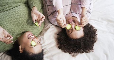 Laughing mother and daughter holding cucumber slices on bed enjoying playful skincare