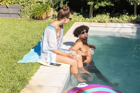 Couple relaxing at pool edge with drinks, smartphone and inflatable float, sunny backyard