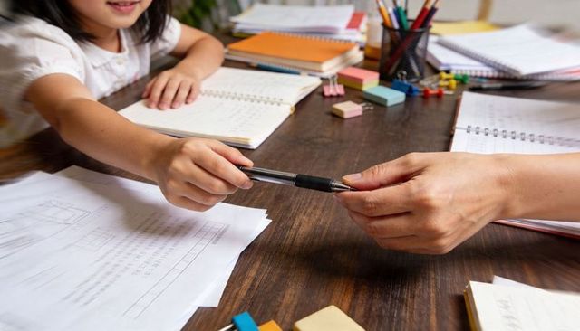 Child passing pen during homework help at wooden desk with notebooks and school supplies