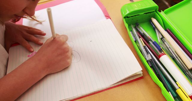 Young school child drawing in a lined notebook, concentrating on their work. Colorful open pencil case full of pens, pencils, and other writing instruments visible. Ideal for use in educational content, back-to-school promotions, creativity and children's learning materials, or stationary advertising.