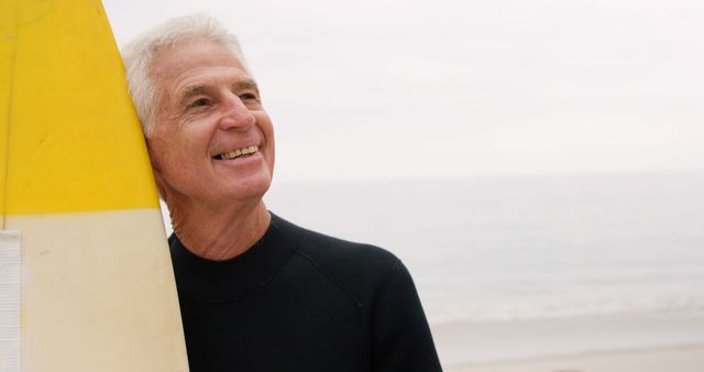 Senior man leaning on surfboard near ocean, looking into distance with a smile. Perfect for illustrating themes of active retirement, healthy lifestyle, water sports, and outdoor recreation. Suitable for magazines, advertisements, blogs, and websites focusing on senior activities, health and wellness, and travel.