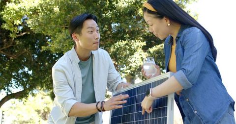 Asian couple examining and installing solar panel outdoors under tree canopy, promoting clean energy