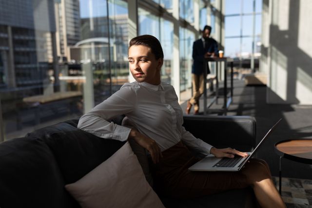 Side view of pretty young Caucasian businesswoman with laptop sitting on the sofa and looking away in modern office. African-American colleague in the background