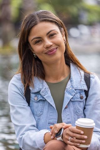 African american woman smiling holding coffee at canal in denim jacket with smartwatch