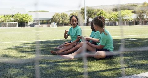 Schoolgirls sitting by goal net on soccer pitch talking and resting during sunny practice