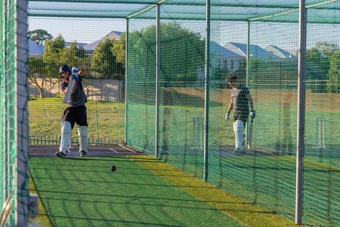 Diverse teammates practicing cricket batting in sunlit outdoor nets with protective pads