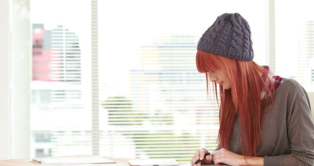 A young Caucasian woman with red hair is focused on her smartphone, sitting indoors with natural light and a cityscape in the background, with copy space. Her casual attire and beanie suggest a relaxed or creative environment.