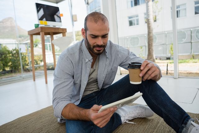 Designer sitting on floor in modern office, holding disposable coffee cup and using tablet. Ideal for themes related to creative workspaces, modern office environments, remote work, and digital technology.