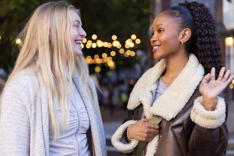 Young diverse women chatting in urban plaza at dusk wearing cardigan and shearling coat