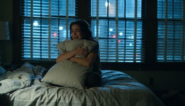 Crying woman clutching pillow on rumpled bed at night under wooden blinds, moody solitude