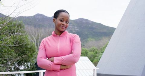African American woman standing with arms crossed on balcony in pink jacket with mountain view