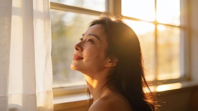 Asian woman gazing through sunlit window during golden hour backlit portrait