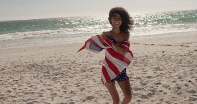 Woman wrapping herself in an American flag, strolling on a sunny beach. Ideal for topics of freedom, summer holidays, patriotism, travel, and carefree lifestyle.