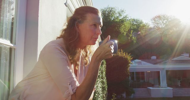 Middle aged woman enjoying a peaceful morning coffee on a balcony. Sunlight gently lighting her side as she gazes into the distance. Ideal for use in lifestyle blogs, promotional materials for wellness products, or articles about morning routines and relaxation.