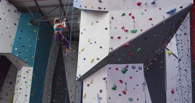 This image shows a woman rock climbing in an indoor gym, equipped with a harness and safety ropes. Colorful climbing holds are visible on the wall, indicating various routes and challenges. This visual is ideal for illustrating concepts related to fitness, sports, hobbies, and active lifestyles. It can be used in promotional materials for gyms, climbing gear companies, or health and fitness blogs.