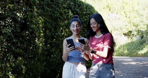 Two Female Friends Smiling While Viewing Smartphones Outdoors
