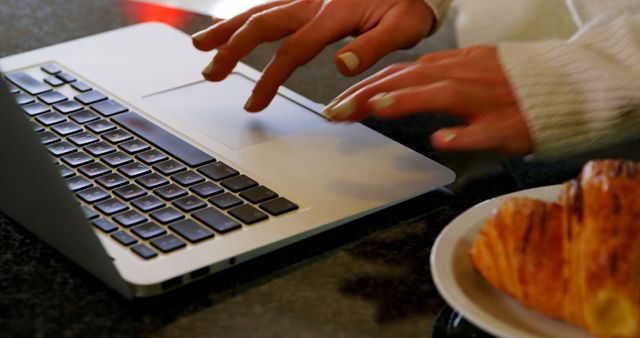This image depicts hands typing on a laptop on a countertop, with a plate of croissant nearby, implying casual work-from-home atmosphere. The cozy kitchen setting suggests a relaxed morning routine. It can be used for articles or blogs about remote working, morning routines, home office setups, or lifestyle.