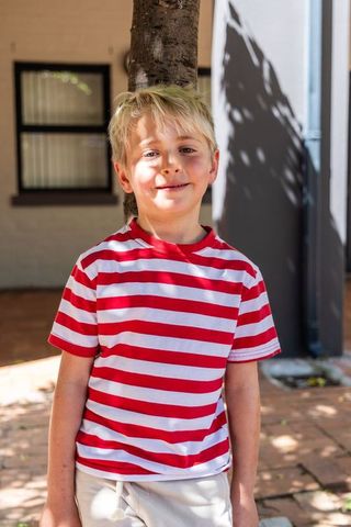 Smiling boy standing by tree on sunlit suburban patio wearing red and white striped t-shirt