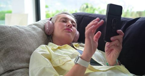 Mature woman reclining on sofa listening with headphones and using smartphone