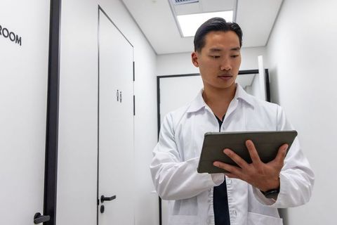 Asian male doctor using tablet in bright clinic corridor, modern healthcare technology