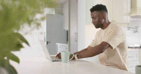 African American man working on laptop at modern bright kitchen island with mint-green mug