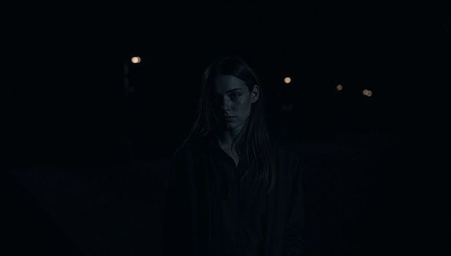 Young woman standing at night in dark hoodie facing camera with streetlight bokeh