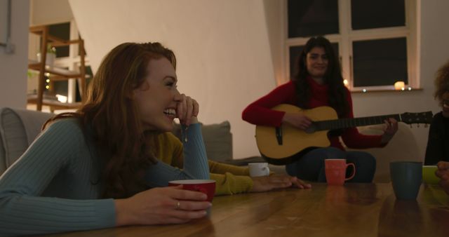 A group of women gathered in a cozy living room, with one playing a guitar while others laugh and drink from mugs. Perfect for themes related to friendship, relaxation, home gatherings, or music. Can be used in articles, blogs, and advertisements focused on home-life, social bonding, or leisure activities.