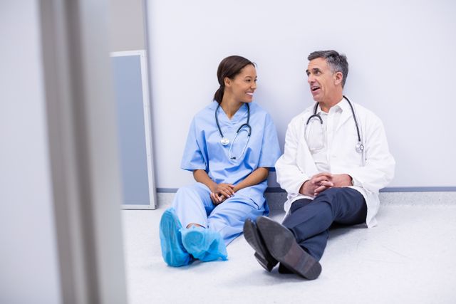 Doctors sitting on floor in hospital corridor, engaging in friendly conversation. Both wearing medical attire, including scrubs and stethoscopes. Ideal for illustrating teamwork, healthcare environment, medical staff interactions, and hospital life. Useful for medical blogs, healthcare websites, and educational materials.