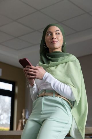 Confident asian woman wearing green hijab and mint pants holding smartphone in office