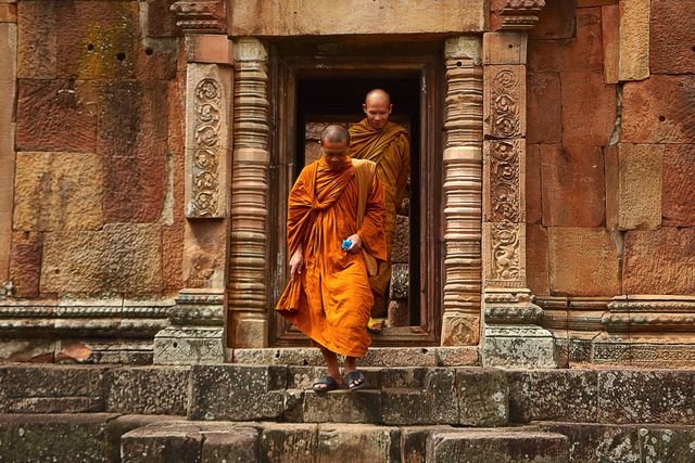 Two Buddhist monks wearing traditional orange robes are leaving an ancient temple in Thailand. The monks are stepping down stone steps, with one monk holding an object in hand. This depiction of religious practice in a historic, cultural setting illustrates spirituality, meditation, and the preservation of cultural heritage. Could be used in content about Southeast Asia travel, cultural studies, religious practices, or historical sites.