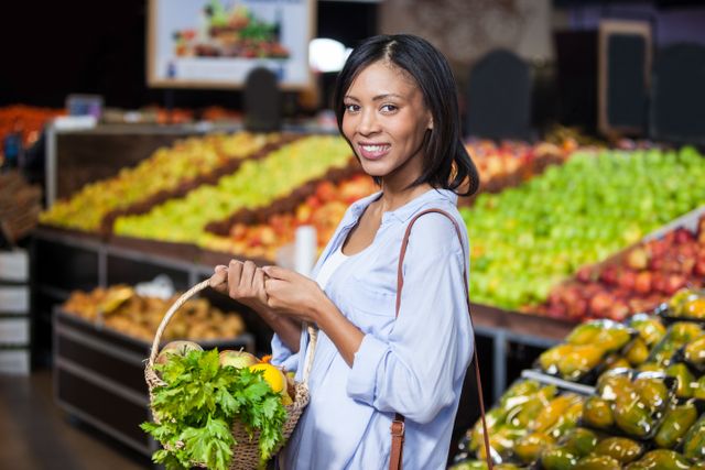 Woman shopping for fresh fruits and vegetables at a supermarket, holding a basket filled with produce. Ideal for use in advertisements for grocery stores, healthy eating campaigns, organic food promotions, and lifestyle blogs.