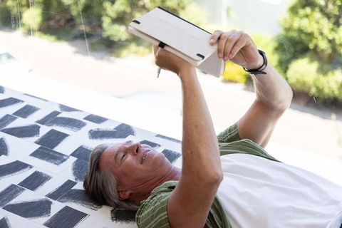 Senior man reclining, reading tablet by sunlit window on patterned mattress, writing notes