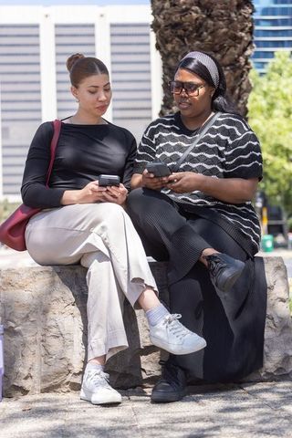 Two african american women sitting on city stone wall using smartphones, sharing messages outdoors
