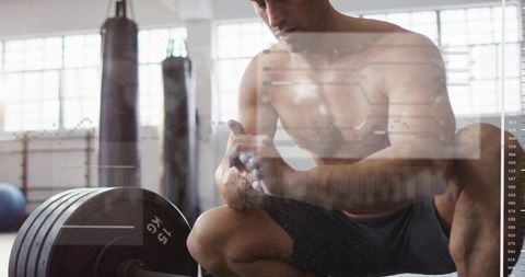 Shirtless athlete applying chalk by loaded barbell in bright industrial gym for strength