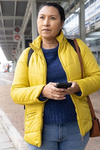 Mature woman standing curbside wearing yellow jacket checking smartphone waiting for pickup