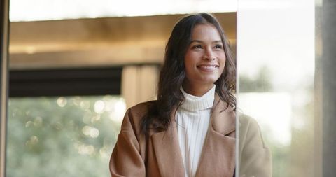 Confident professional woman in camel coat smiling while looking out large office window