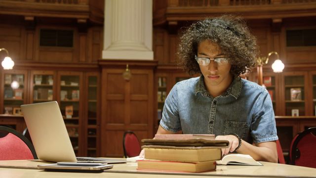 Young man focused on studying in a historic library, using books and a laptop. Ideal for educational content, academic study illustration, or promoting library environments. It evokes a sense of concentration and scholarly atmosphere.